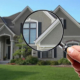 A hand holding a magnifying glass in front of a suburban home to signify an inspection